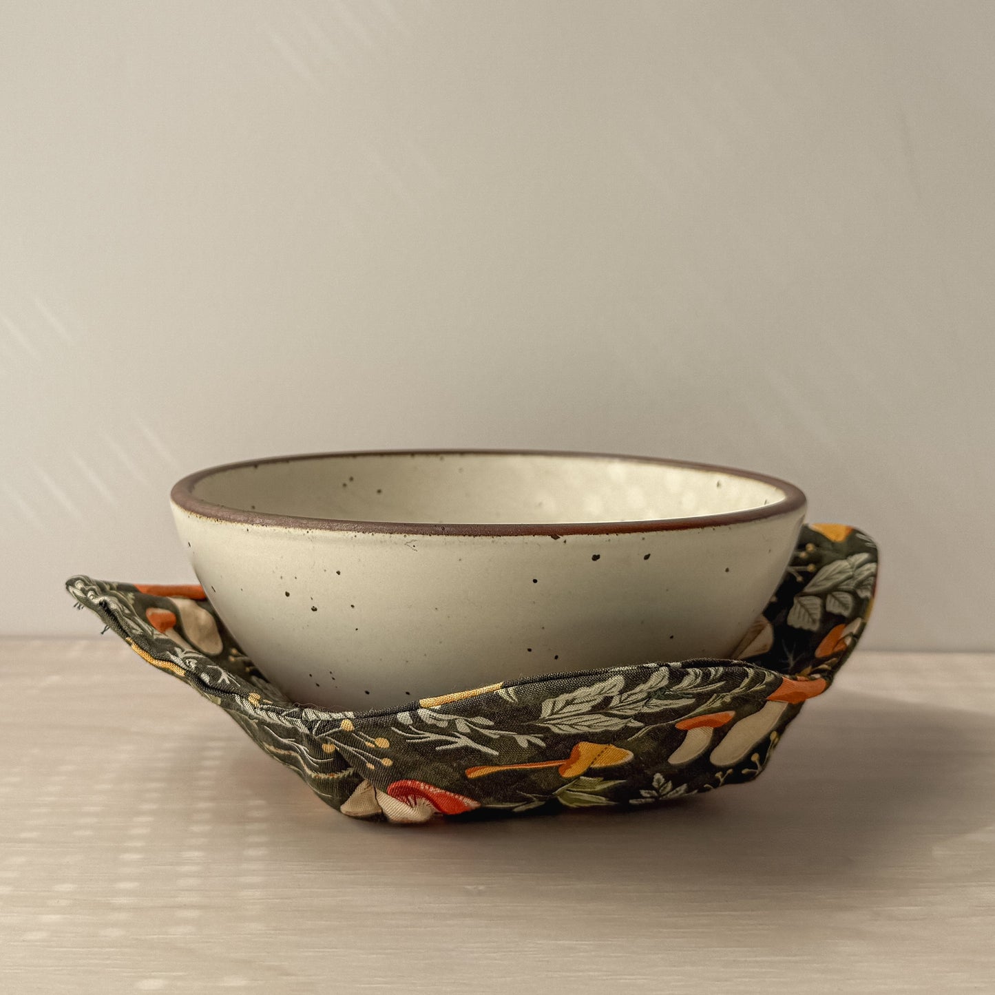 Ceramic bowl cushioned inside a mushroom patterned bowl cozy on a neutral background