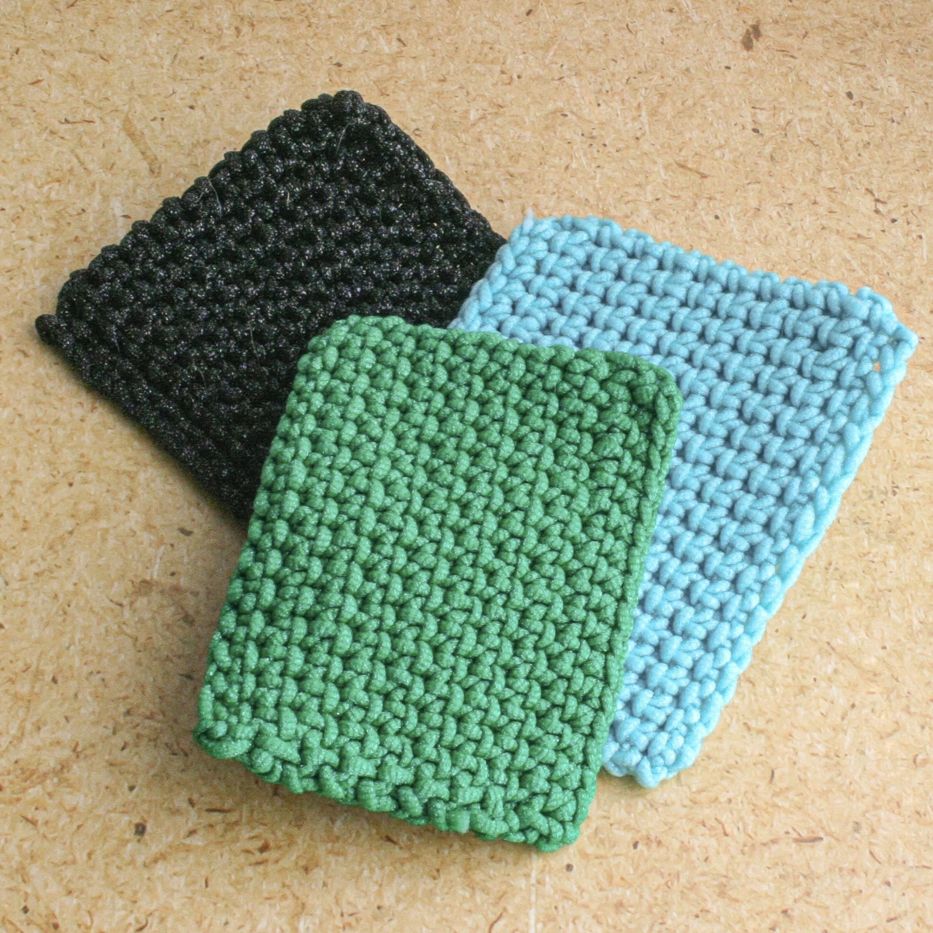 Three crocheted dish Scrubbies in black, green, and blue on a beige background