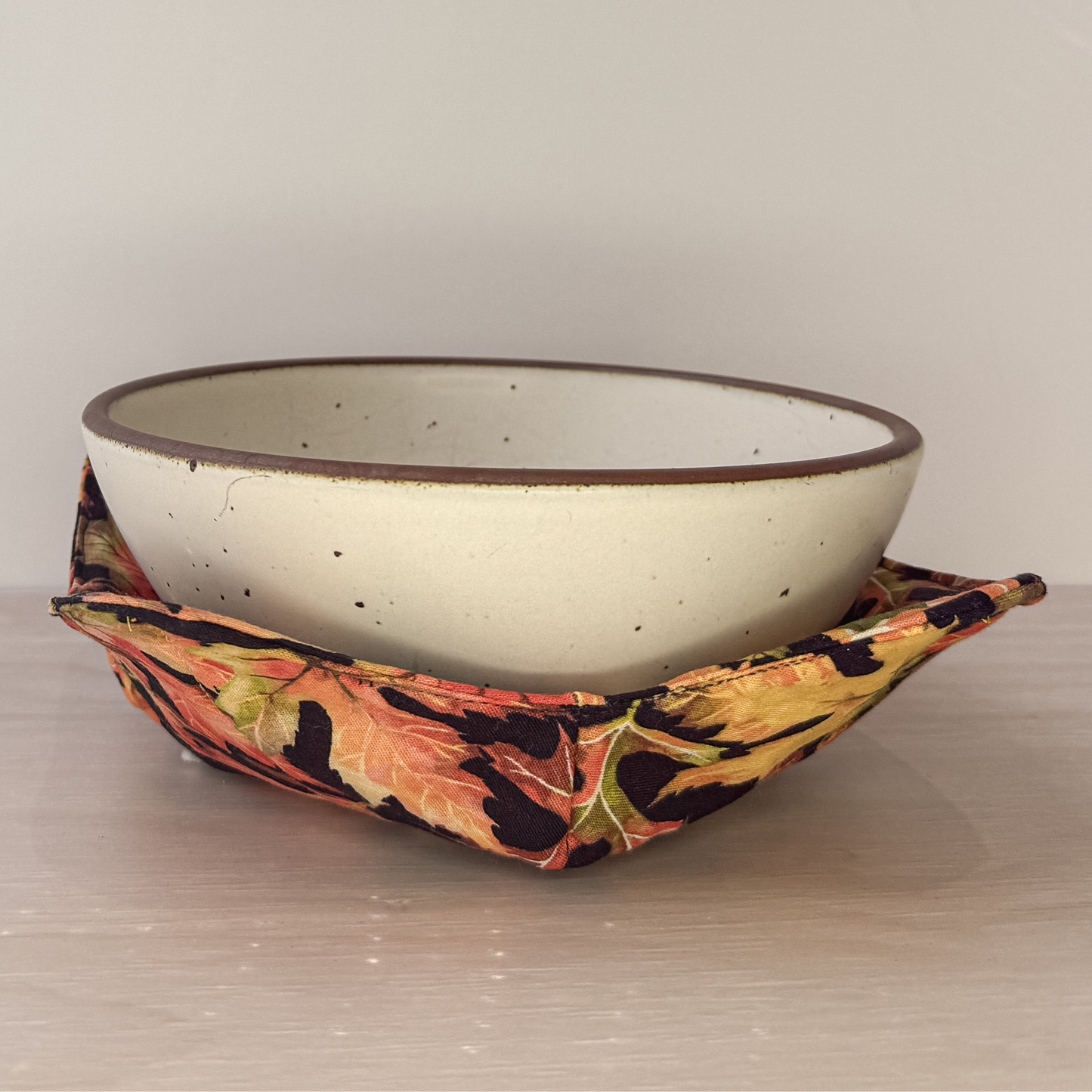 An off white bowl nestled in a bowl cozy. The bowl cozy is a combination of orange red and gold leaves on a black background