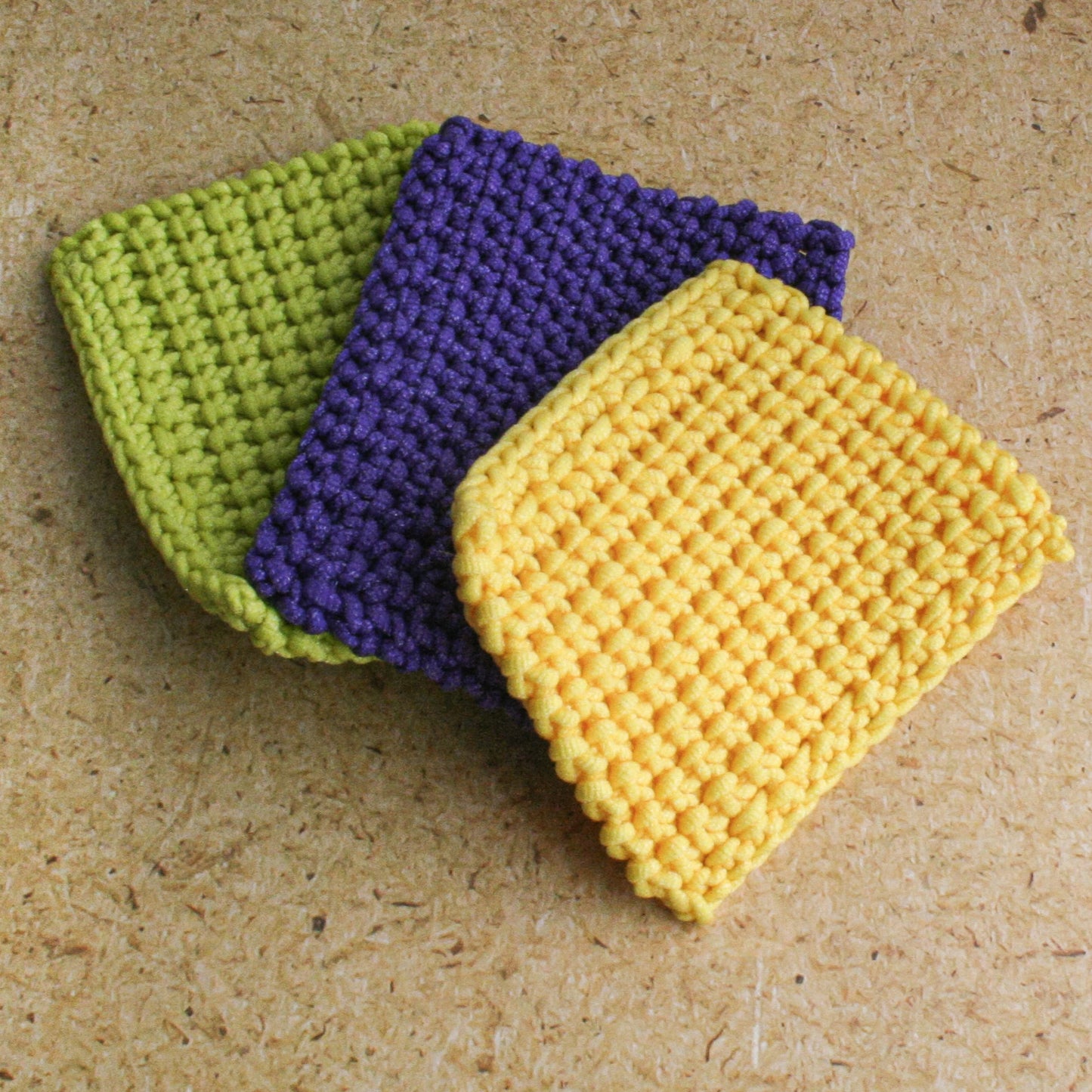 Three crocheted Scrubbies in lime green, purple, and yellow on a textured surface.