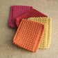 Four Crocheted squares in pink, red, orange, and yellow on a textured surface.